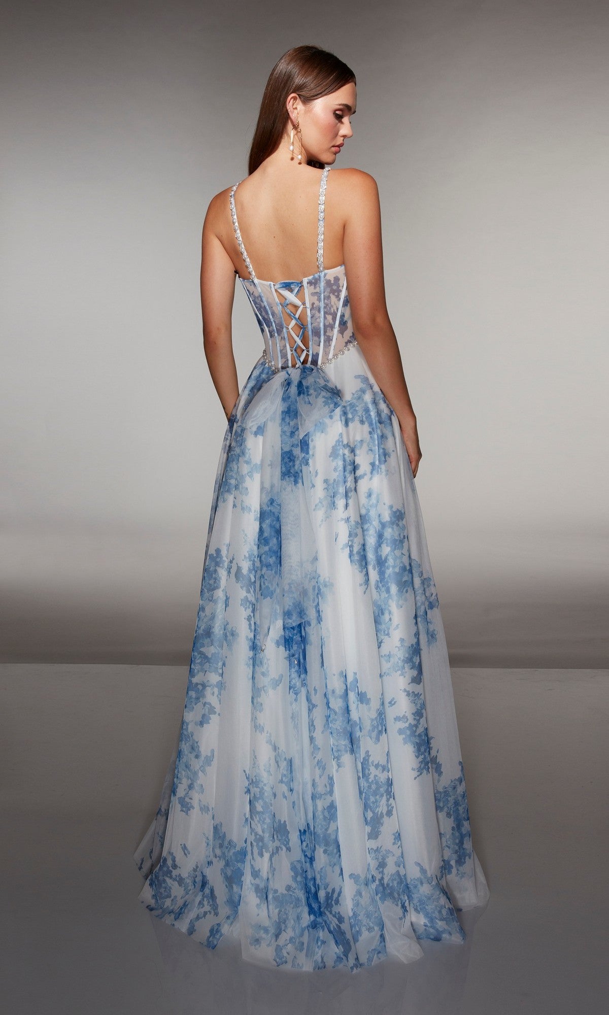 Woman in a floor-length, sleeveless Alyce 61994 prom dress with blue floral patterns and a lace-up corset bodice, standing and facing away from the camera.